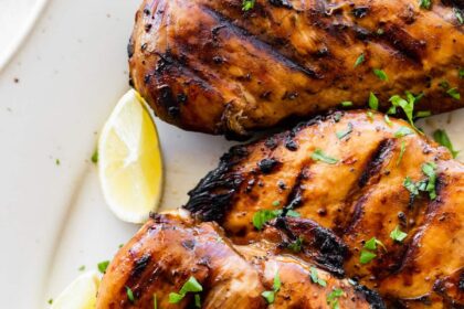 Four marinated grilled chicken breasts on a plate topped with fresh herbs and lemon wedges on the side.