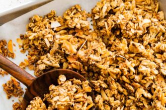 A baking sheet with a large wooden spoon taking a portion of coconut granola.