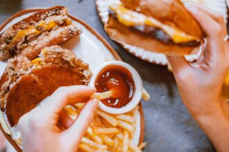 Image of a person dipping a French fry in ketchup with one hand and holding a cheeseburger in the other hand.