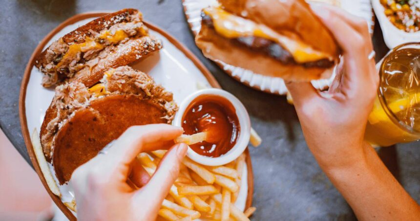 Image of a person dipping a French fry in ketchup with one hand and holding a cheeseburger in the other hand.