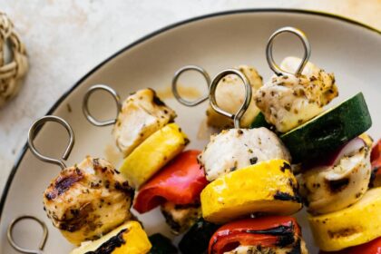 Six grilled chicken kabobs on a white plate. The kabobs combine chicken, yellow squash, zucchini, red pepper and onion.