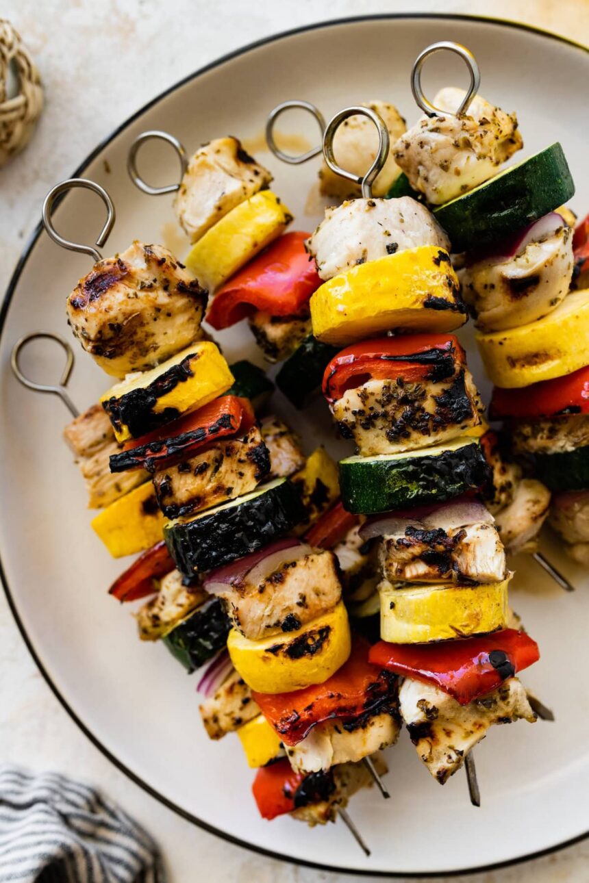 Six grilled chicken kabobs on a white plate. The kabobs combine chicken, yellow squash, zucchini, red pepper and onion.