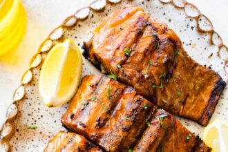 Four grilled salmon filets on a large plate with lemon wedges on the side.