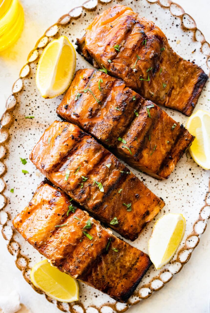 Four grilled salmon filets on a large plate with lemon wedges on the side.