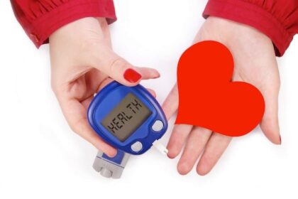 Two female hands with the left holding a blood glucose meter and the right holding a red cardboard heart.