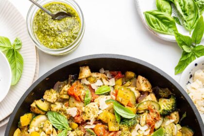 A large skillet of cooked pesto chicken with multiple vegetables and topped with fresh basil leaves.