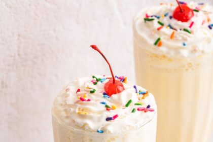 A protein milkshake in a large glass cup topped with whipped cream, sprinkles, and a cherry.