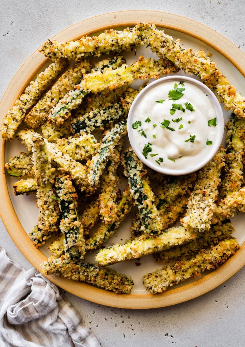 A large plate full of crispy zucchini fries covered in panko bread crumbs with a small dish of a dip.