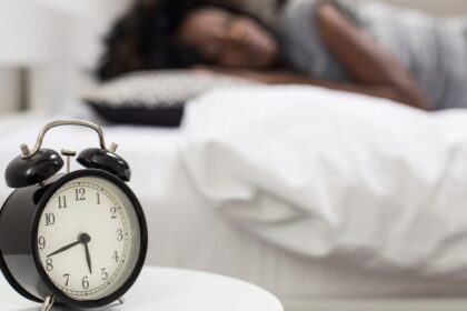 Close-up photo of an alarm clock and a woman sleeping in the background.