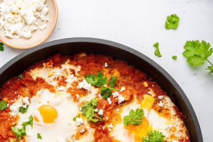 A cast iron skillet with shakshuka. It contains poached eggs in a tomato based sauce and is topped with crumbled feta cheese and fresh herbs.