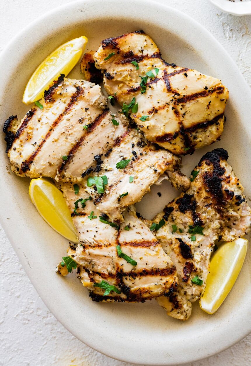 Multiple yogurt marinated chicken thighs on a plate with lemon wedges on the side.