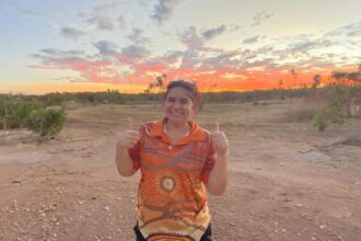 Aboriginal woman with her hair in a pigtail and wearing an orange Aboriginal shirt stands in a desert at sundown giving a double thumbs up.