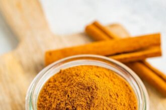 Small glass jar with pumpkin pie spice on a wood board and next to two sticks of cinnamon.