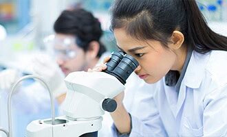 A young female researcher is looking into a double-lens microscope.