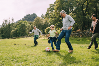 Older man with grey hair kicks a ball in a green field with kids and a young adult.