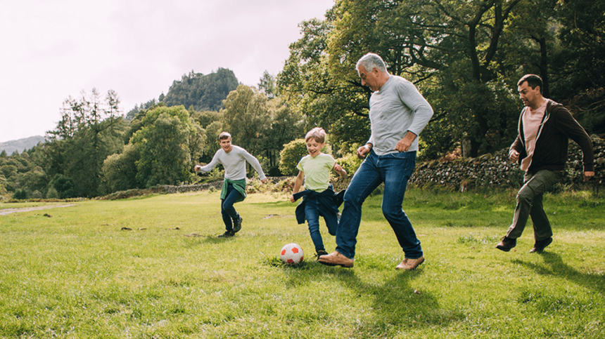 Older man with grey hair kicks a ball in a green field with kids and a young adult.