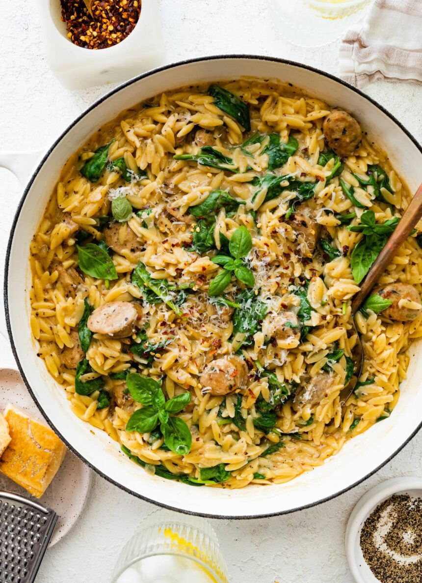 A white Dutch oven with creamy orzo, chicken sausage and spinach mixed together. A brown serving spoon is in the pot on the right hand side.