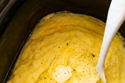 Mashed potatoes in a slow cooker with slices of butter, seasonings, and a spatula.