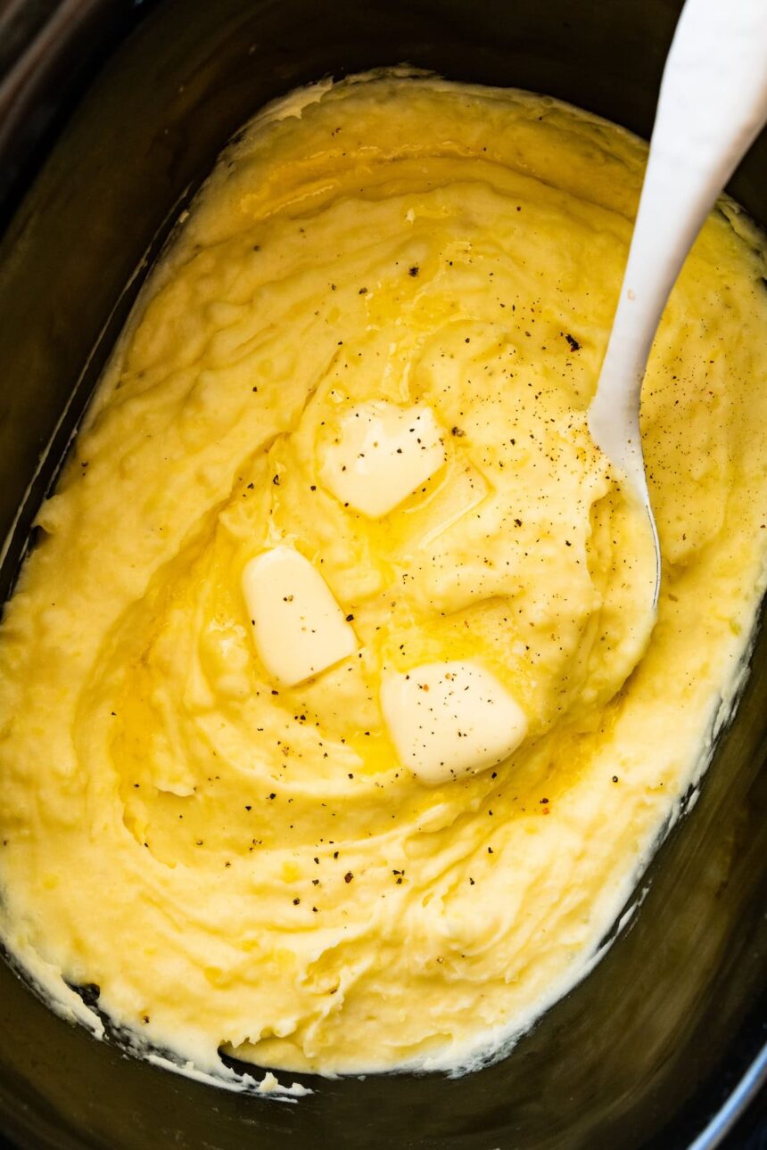 Mashed potatoes in a slow cooker with slices of butter, seasonings, and a spatula.