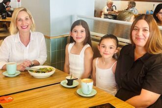 Justine Cain, Olivia with straight, long brown hair parted in the middle, Emily, with her hair pulled off her face in a tight ponytail, and their Mum, Yvette, sit at a booth at a coffee shop with their drinks and food on a wooden table.