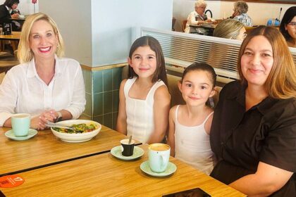 Justine Cain, Olivia with straight, long brown hair parted in the middle, Emily, with her hair pulled off her face in a tight ponytail, and their Mum, Yvette, sit at a booth at a coffee shop with their drinks and food on a wooden table.