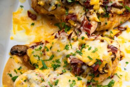 Four chicken breasts in a white baking dish topped with mushrooms, turkey bacon, melted cheese and fresh parsley.