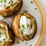 Three baked potatoes split open on a plate with sour cream, fresh chives, and seasoning.