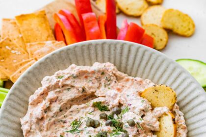 A bowl of smoked salmon dip, garnished with fresh dill and capers, served with crackers and fresh vegetables.