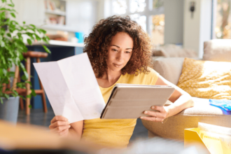 Woman with light brown curly hair to her shoulders holds a sheet of paper in one hand and compares the information on the tablet she is holding while sitting on a sofa.