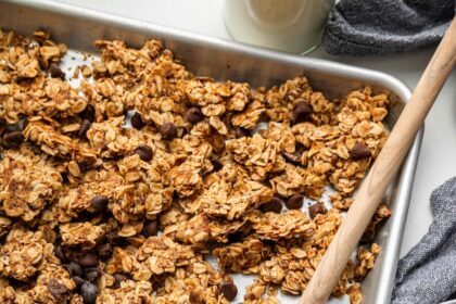 Clusters of chocolate chip cookie granola on a silver baking sheet served with a wooden spoon.