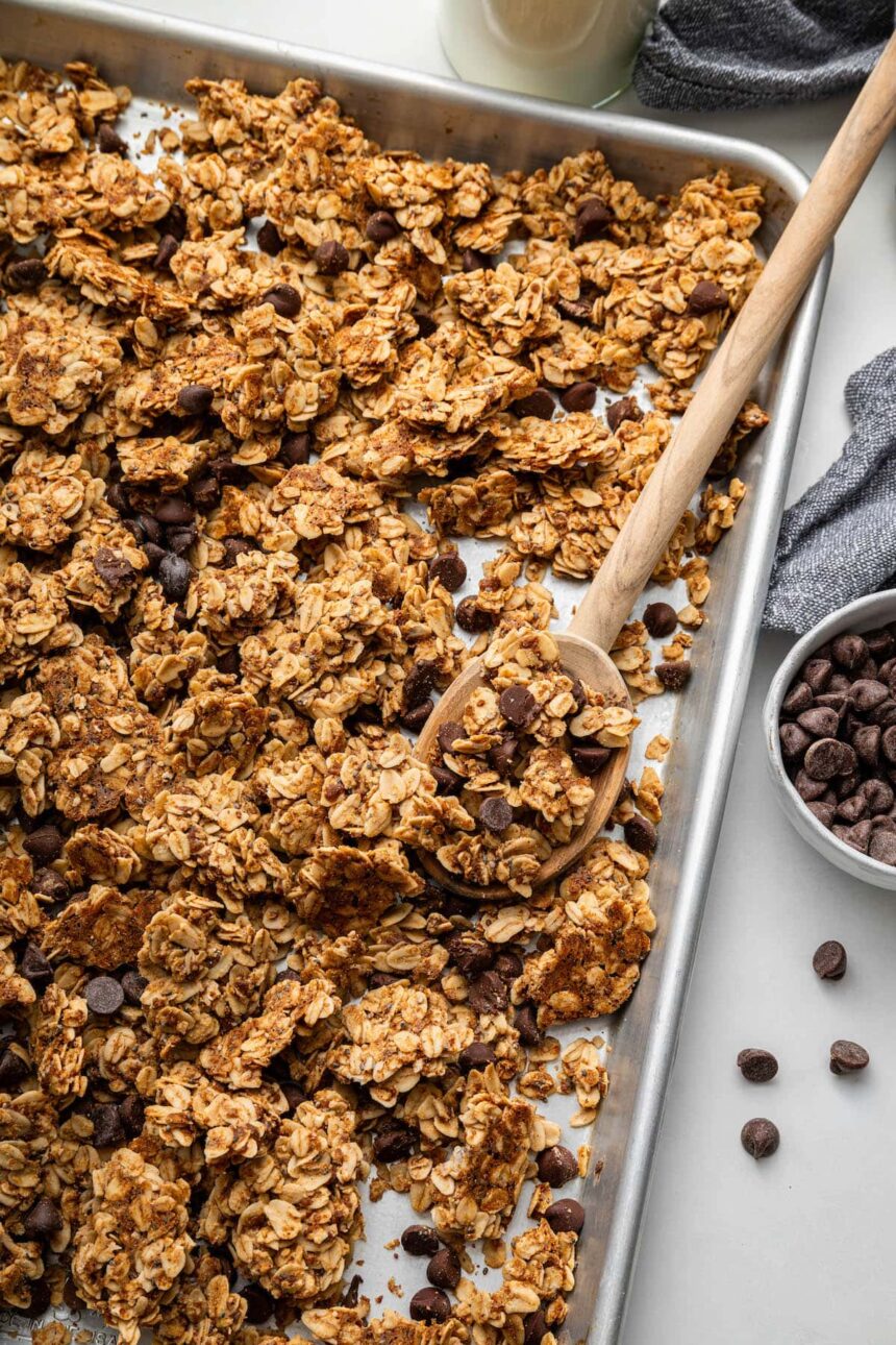 Clusters of chocolate chip cookie granola on a silver baking sheet served with a wooden spoon.