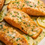 Four salmon filets in a pan with garlic butter and lemon wedges. The filets are garnished with fresh herbs.