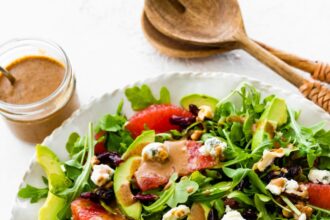 A salad with arugula, avocado, blue cheese, grapefruit, nuts, and cranberries on a white plate.