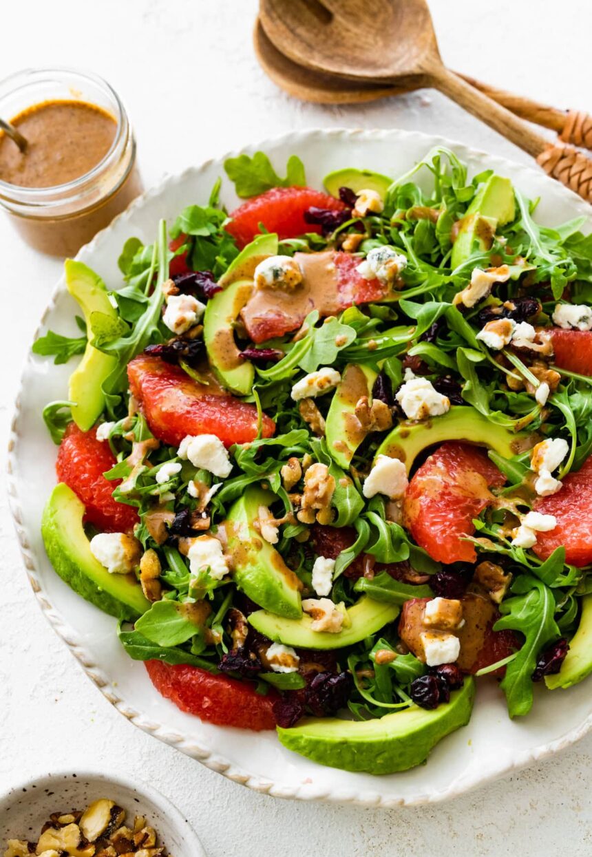 A salad with arugula, avocado, blue cheese, grapefruit, nuts, and cranberries on a white plate.