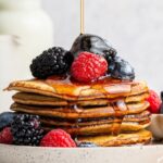 Maple syrup being poured on top of a stack of six greek yogurt pancakes on a plate. They