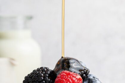 Maple syrup being poured on top of a stack of six greek yogurt pancakes on a plate. They