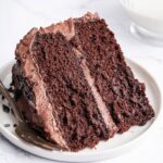 A slice of chocolate cake with chocolate buttercream frosting on a white plate with a metal fork to the side.