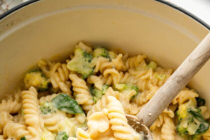 Rotini pasta, broccoli and spinach in a pot coated in a cheese sauce. A wooden spoon is scooping up some of the pasta.