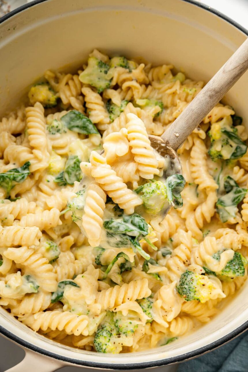 Rotini pasta, broccoli and spinach in a pot coated in a cheese sauce. A wooden spoon is scooping up some of the pasta.