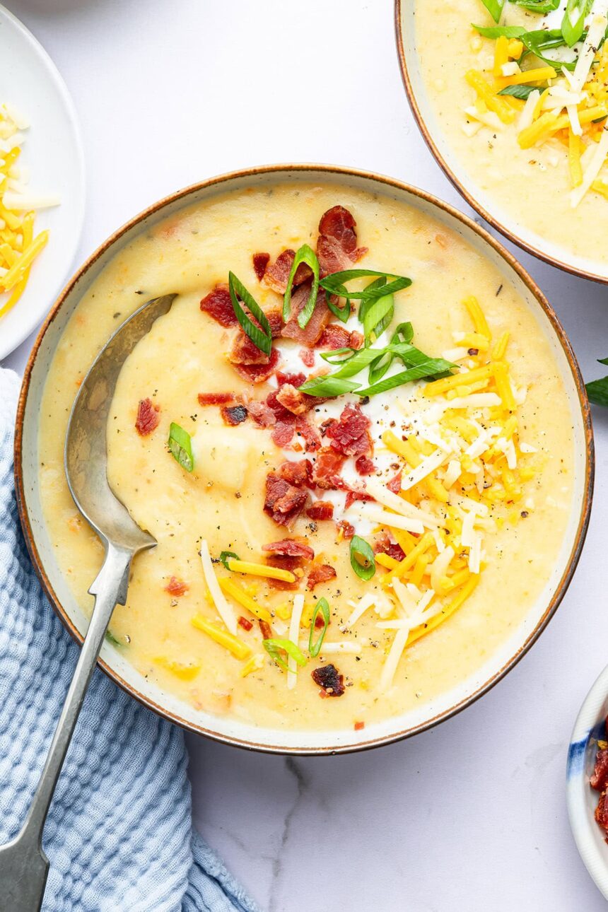 A bowl of potato soup with a silver spoon in it. The soup is topped with bacon bits, green onion, shredded cheese and sour cream.