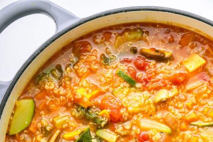 A large pot of vegetable packed quinoa soup with a wooden serving spoon in the soup.