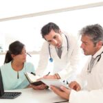 Female in a green top, with a man in a white coat, and another man standing between them looking at patient files.