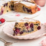 A slice of mixed berry cake on a light pink plate. The entire berry cake is in the background on a white plate.