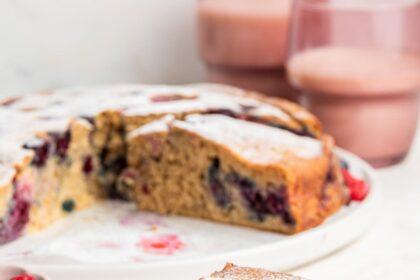 A slice of mixed berry cake on a light pink plate. The entire berry cake is in the background on a white plate.