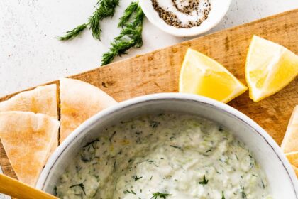 A white bowl of tzatziki topped with fresh dill. There is a wooden spoon in the bowl and the bowl is surrounded by pita bread and lemon slices.