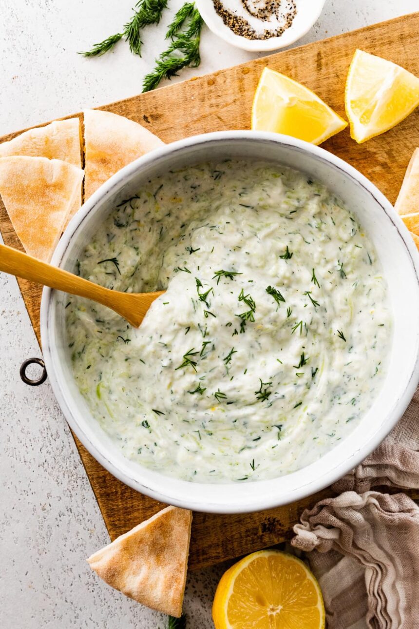 A white bowl of tzatziki topped with fresh dill. There is a wooden spoon in the bowl and the bowl is surrounded by pita bread and lemon slices.