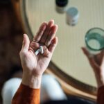 Overhead picture of a woman holding pills in one hand and a glass of water in the other with other medications on a table under her hand.