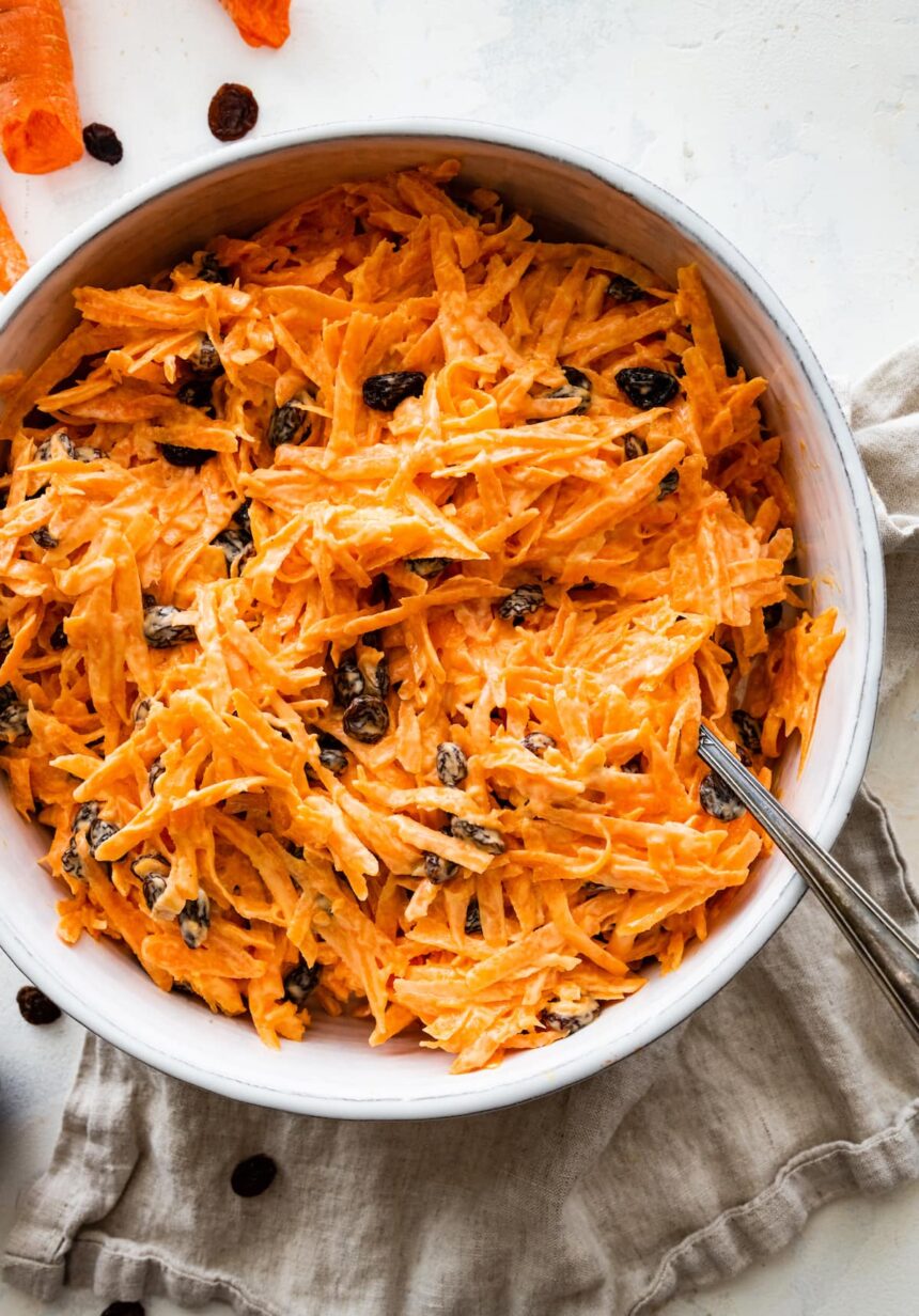 A carrot raisin salad in a white bowl with a spoon.