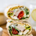 Two halves of a Greek chicken wrap stacked on top of each other on a wooden cutting board. You can see the tomatoes, chicken, lettuce and sauce inside the wrap.