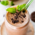 One mint chocolate protein shake in a glass topped with crushed Thin Mint cookies and fresh mint. There is a green straw in the glass.